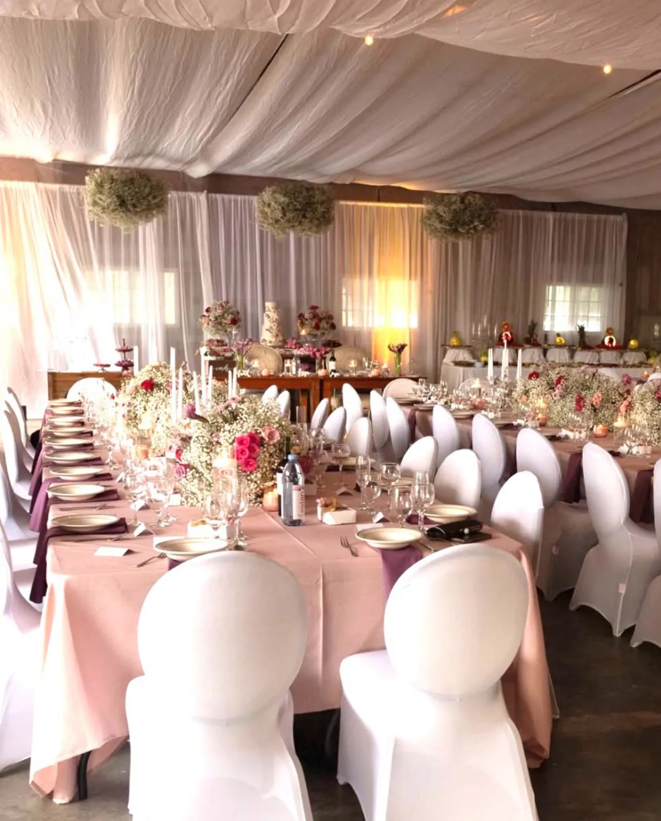 A barn reception set for dinner — long tables in blush linen with candles, deep red roses and baby's breath.