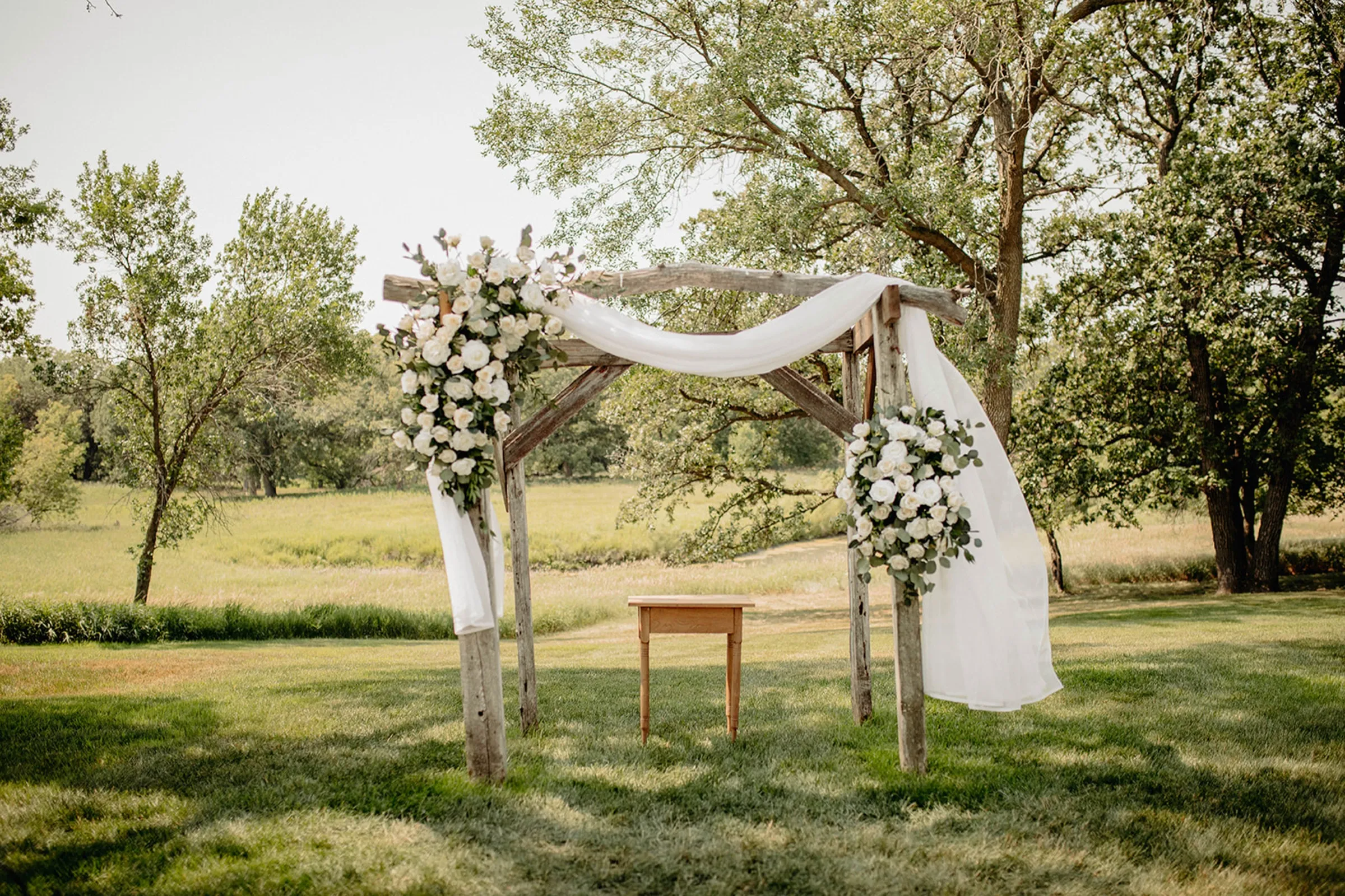 A wooden ceremony arch dressed with white florals and draped fabric, standing in an open field ringed by trees.