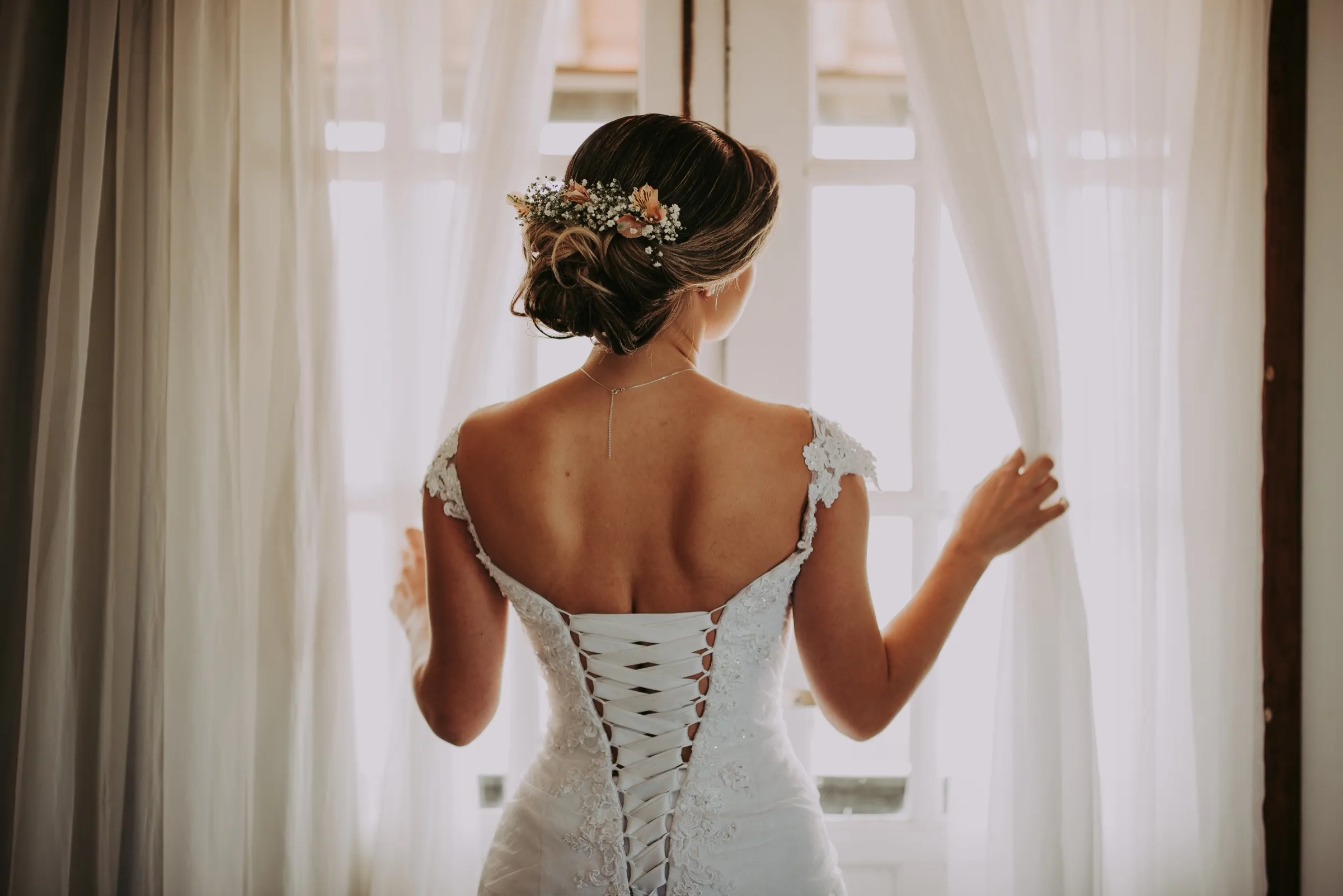 A bride at a window in soft morning light, the corseted back of her gown and floral hairpiece in detail.